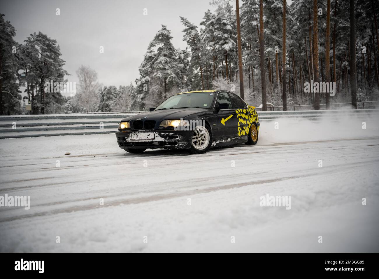 12-12-2022 Riga, Latvia  a black car driving down a snow covered road next to trees and a fence with yellow stickers on it. . Stock Photo