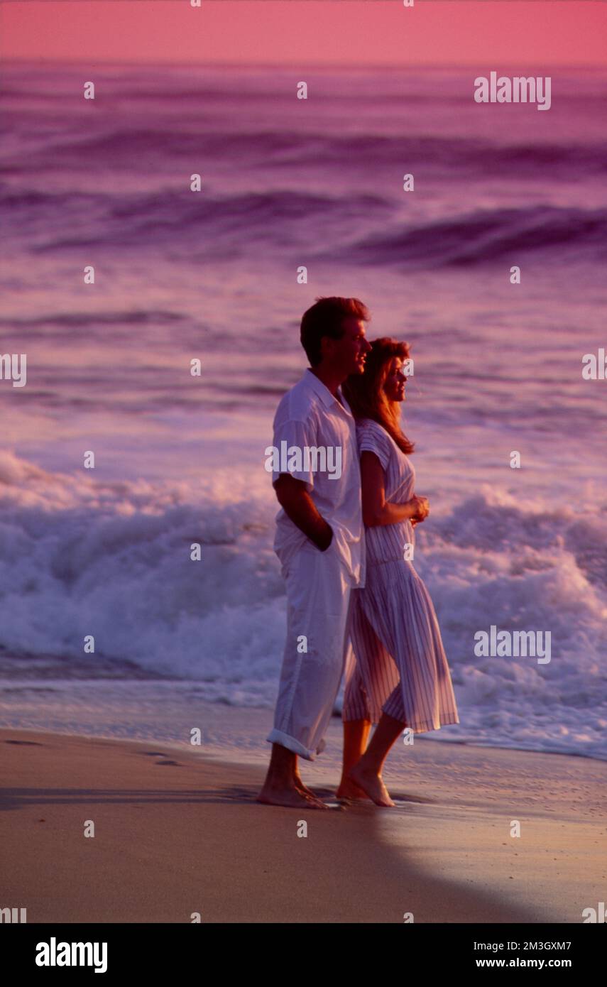 Young couple in white on romantic walk along the beach at sunset Stock Photo