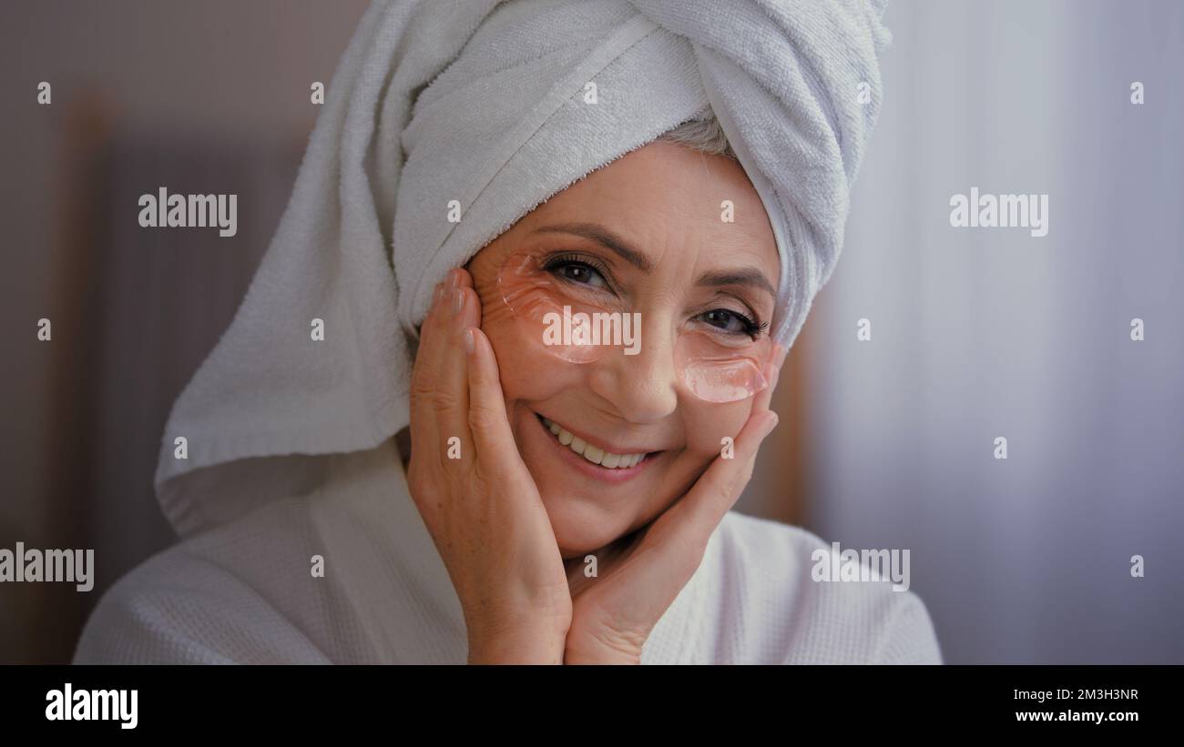 Positive portrait happy senior lady with anti wrinkle skin hydrogel collagen patches under eyes smiling to camera old 60s Caucasian woman with towel Stock Photo