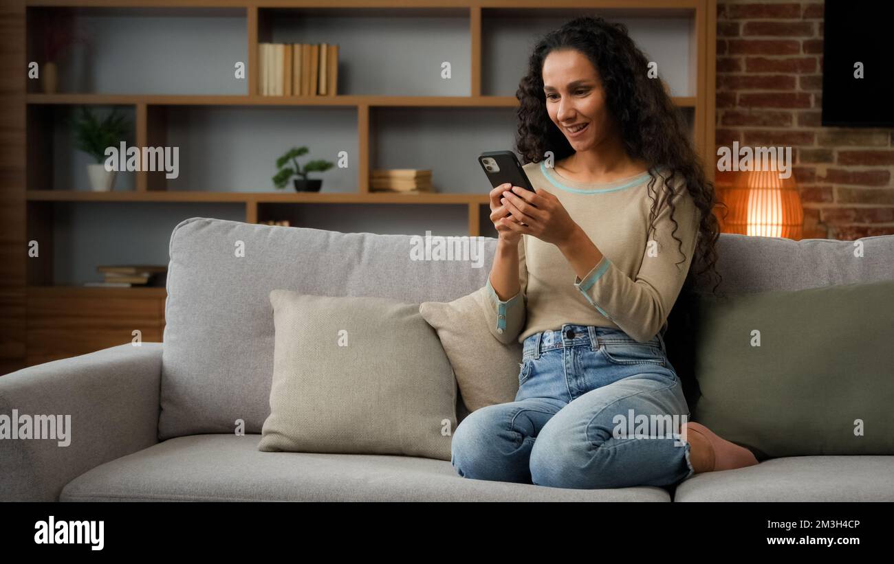 Adult woman lady using phone for bet in internet receive message with good news offer sitting on couch at home win mobile phone win game online Stock Photo
