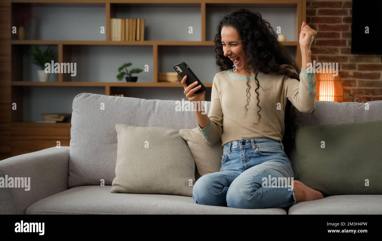 Happy woman lady using phone for bet in internet receive message with good news offer sitting on couch at home win mobile phone win game online Stock Photo