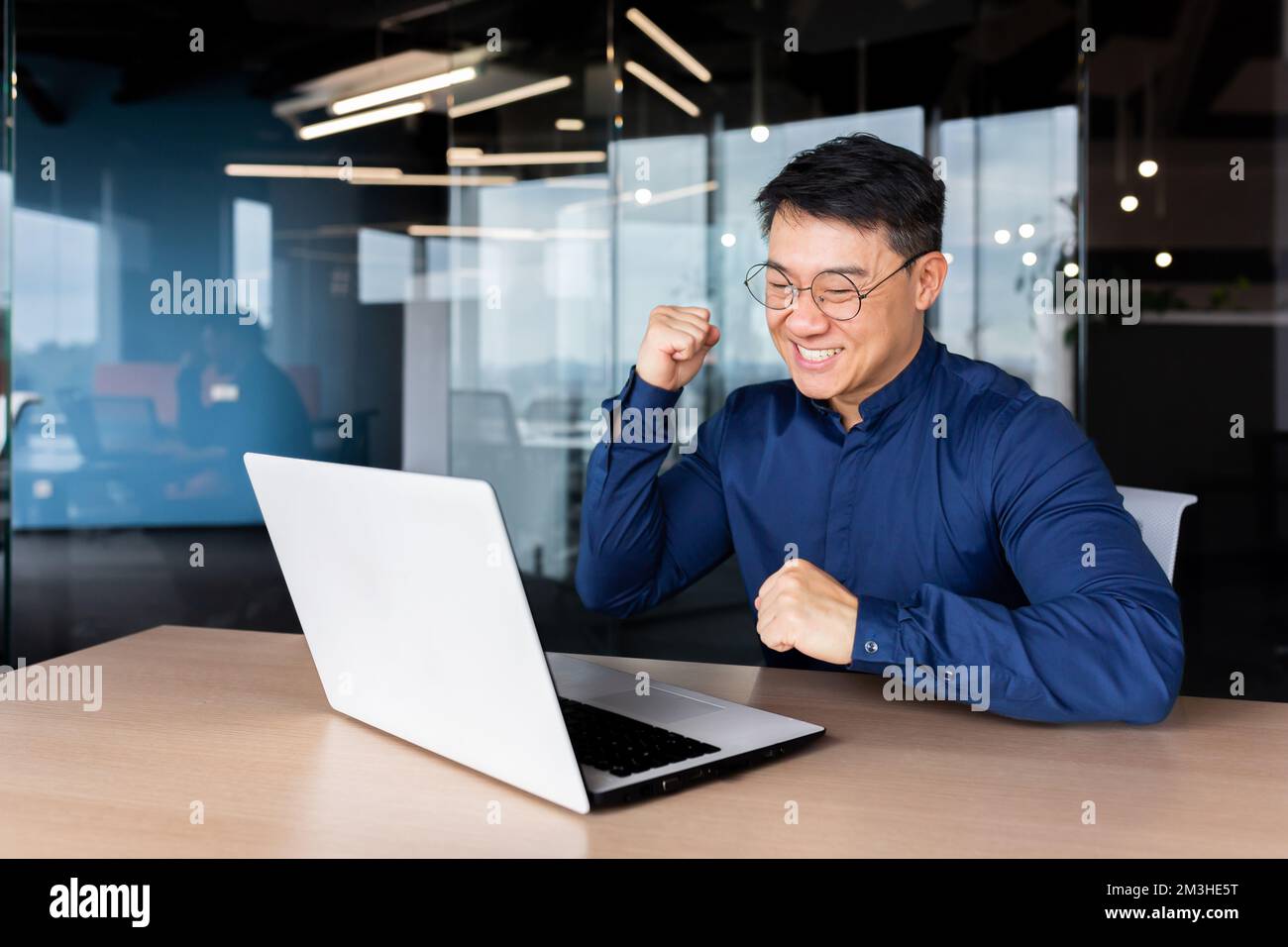Successful asian man celebrating victory and success received news of good achievement online, businessman looking at laptop screen and holding hand up triumph gesture, worker inside office. Stock Photo