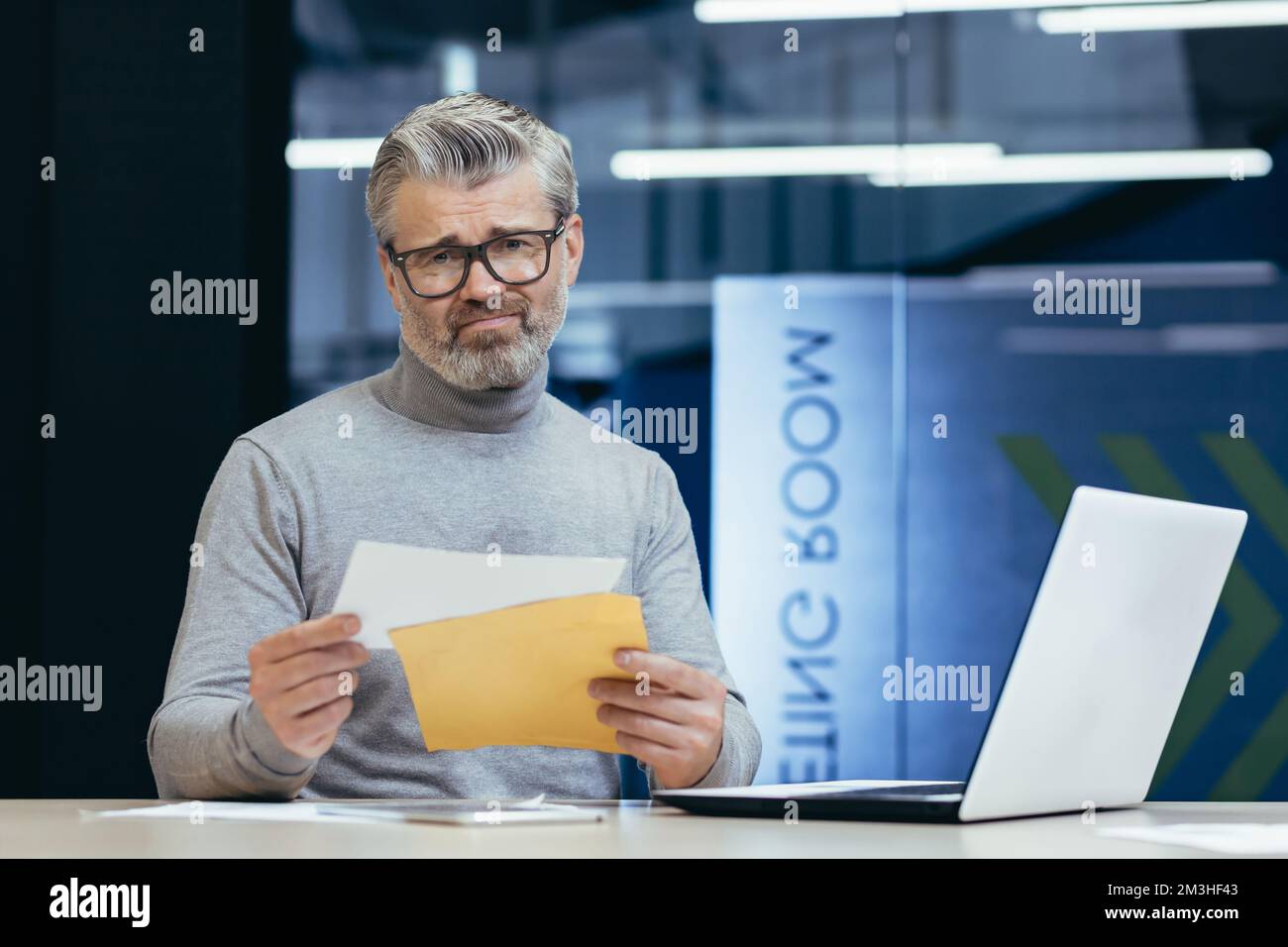 Upset senior man businessman sitting in the office at the desk, holding an envelope with a letter in his hands, received bad news, bank account for a loan, debt. Stock Photo