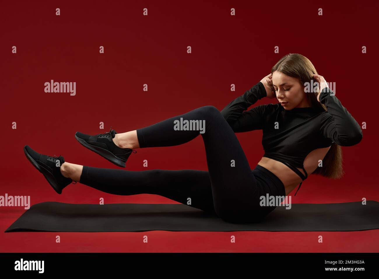 Pretty brunette woman trainer doing twists and abs sitting on yoga mat. Female in black sports suite training cross fit exercise on red studio background. Concept of cardio training. Stock Photo