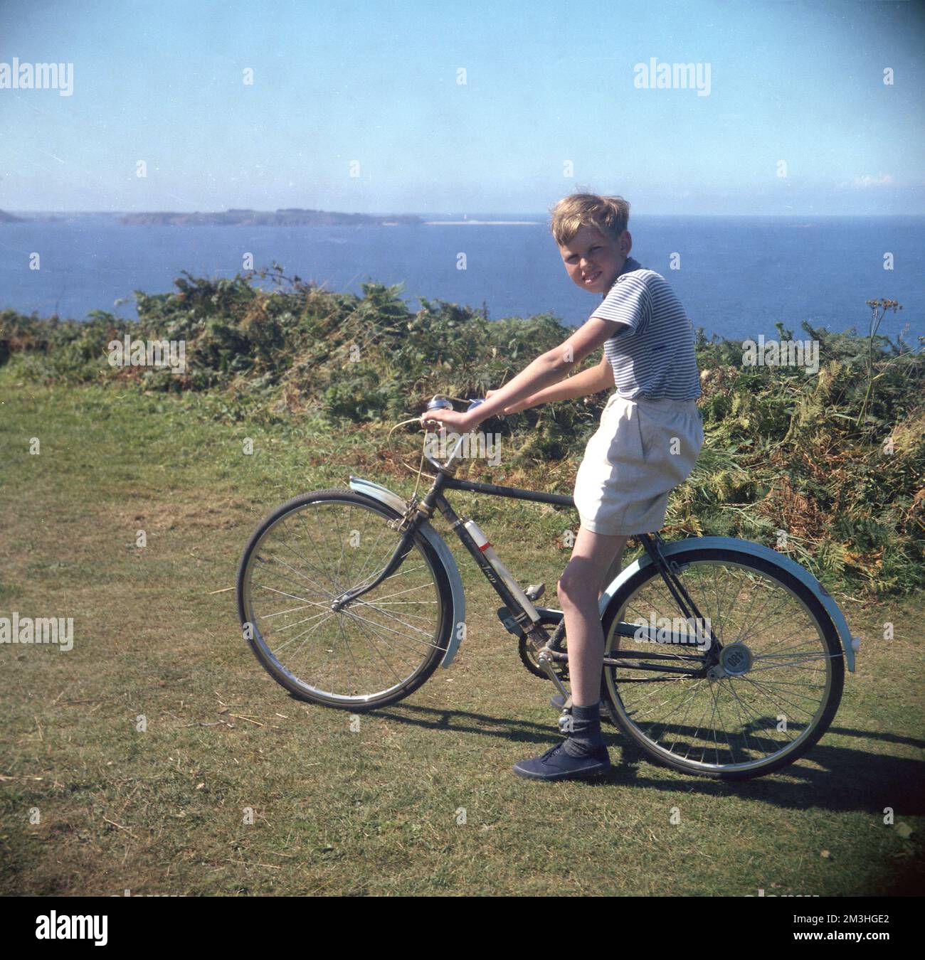1960s, historical, a teenage boy on his bicycle of the era, a Hercules Jeep, on a grassy path overlooking the coast, England, UK, The youngster, wearing a short-sleeve striped top, shorts and blue plimsolls, is sitting astride the British mini-roadster, which has a bicycle pump on the frame. Stock Photo