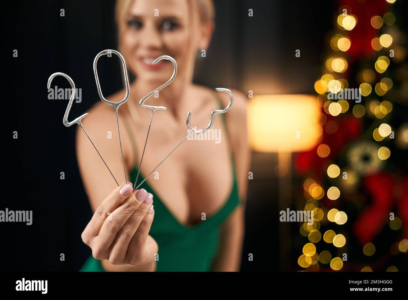 Front view of blonde woman holding 2023 sparkles, showing. Pretty, cheerful lady wearing green, seductive dress, looking at camera, smiling. Concept of New Year and Christmas. Stock Photo