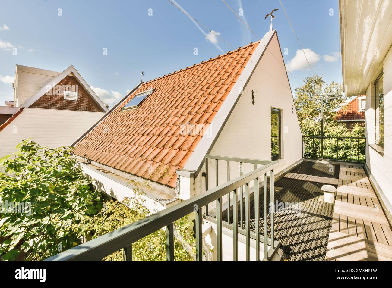 a balcony with an orange tiled roof and white sidings on the house is surrounded by lush green trees in the background Stock Photo