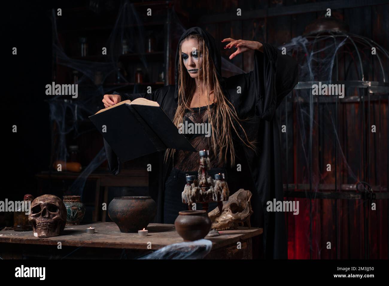 Witch dressed dark dungeon room use magic book for conjuring magic spell. Female necromancer wizard gothic interior with skull, cage, spider web Stock Photo