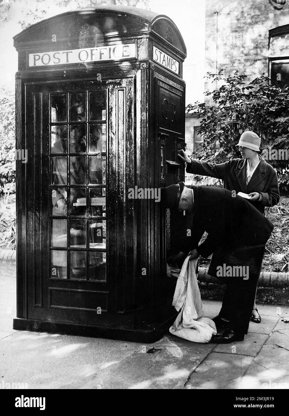 A new telephone kiosk in use in a London street, showing a caller inside, postage stamps being bought from the machine outside, and a postman collecting mail from the letterbox.  1928 Stock Photo