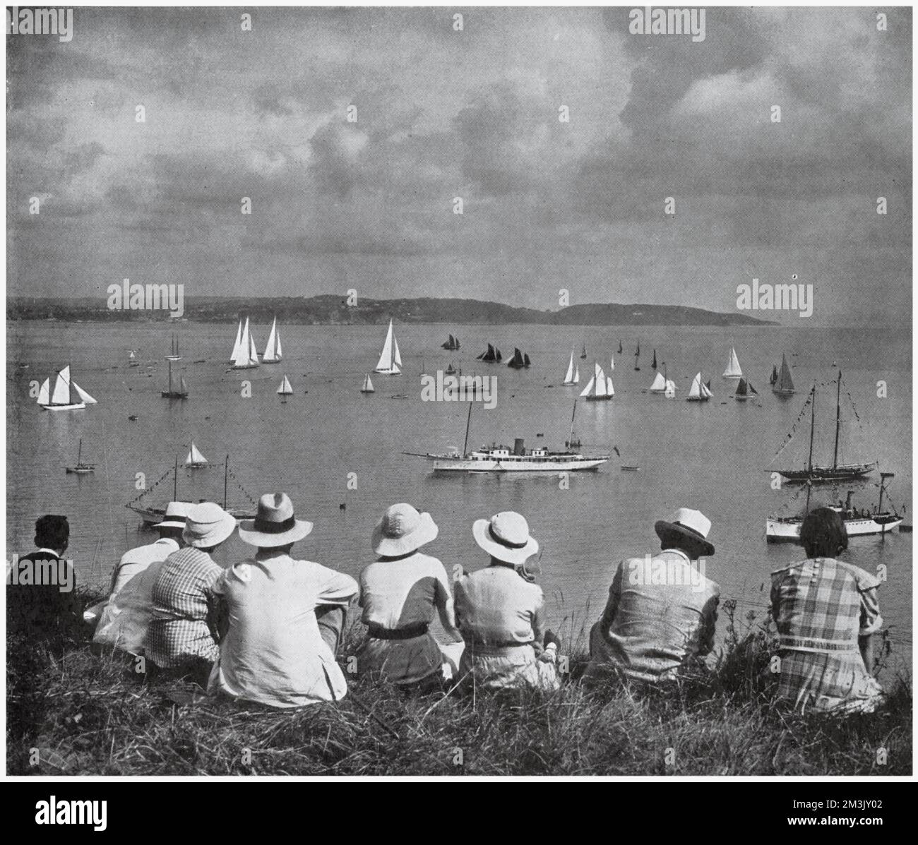 A group of holidaymakers watching the Brixham Regatta, from a vantage point on a West Country cliff, 1936. A number of fishing trawlers, with dark sails, share the waters with the white sails of racing yachts. Stock Photo