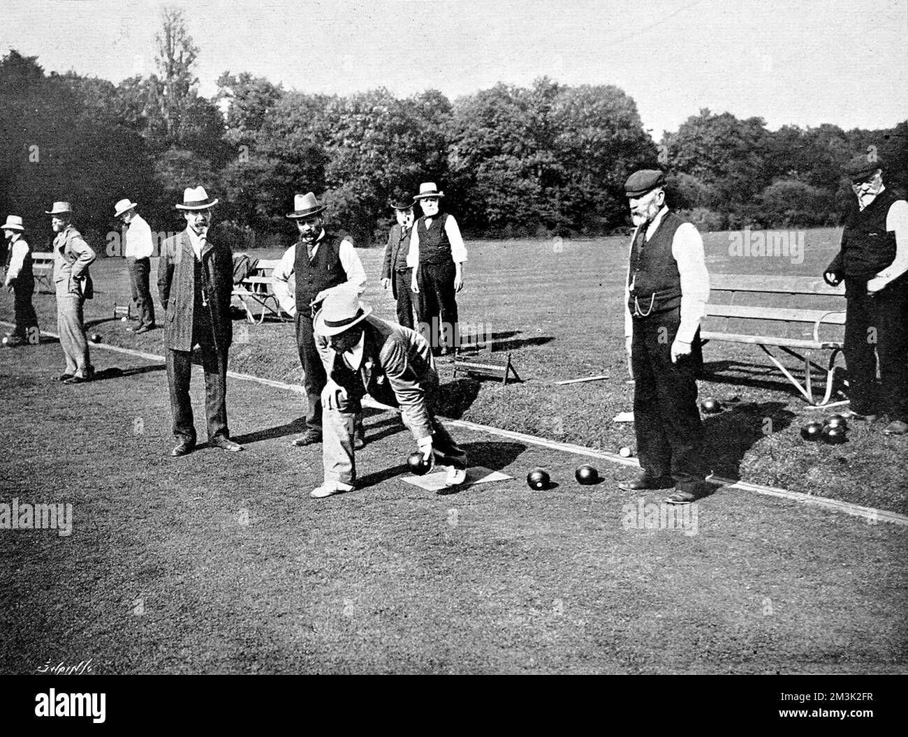 An English gentleman about to deliver his bowl during a game of lawn bowling. Fellow players and spectators closely watch his technique. Stock Photo