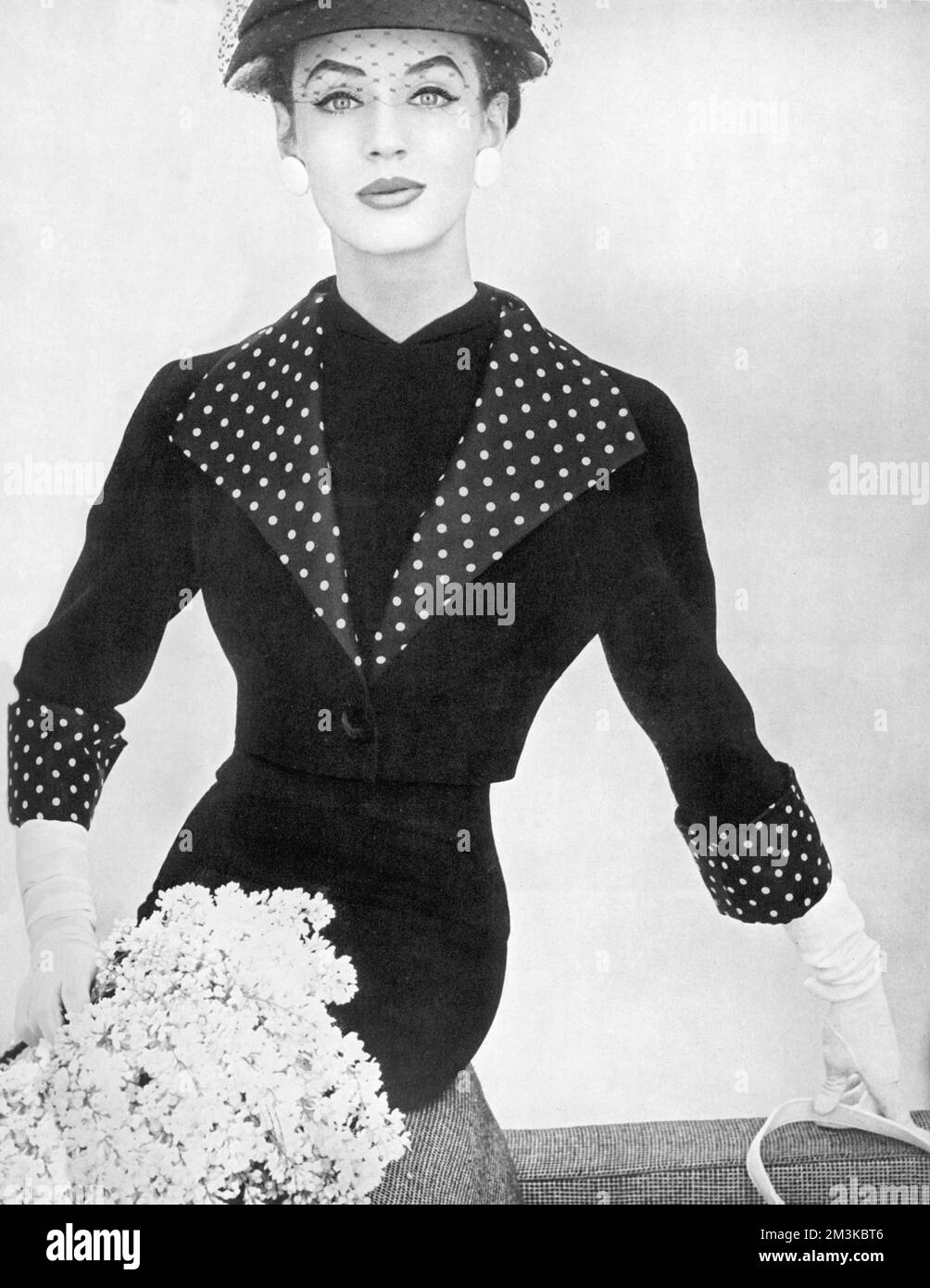 A bolero and dress of lightweight wool georgette with all accessories from Debenham and Freebody, is a spring winner.  The bolero, high lighted with a spotted silk surah and the dress beneath with the straight long look cost 20gns. The hat is a navy shantung Baku cloche.     Date: 1956 Stock Photo