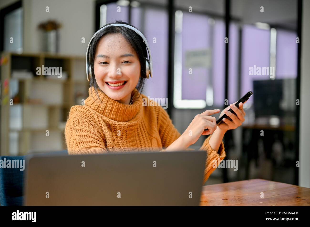 Cheerful and pretty young Asian female looking at laptop screen, holding her smartphone and listening to music through her headphones. Stock Photo