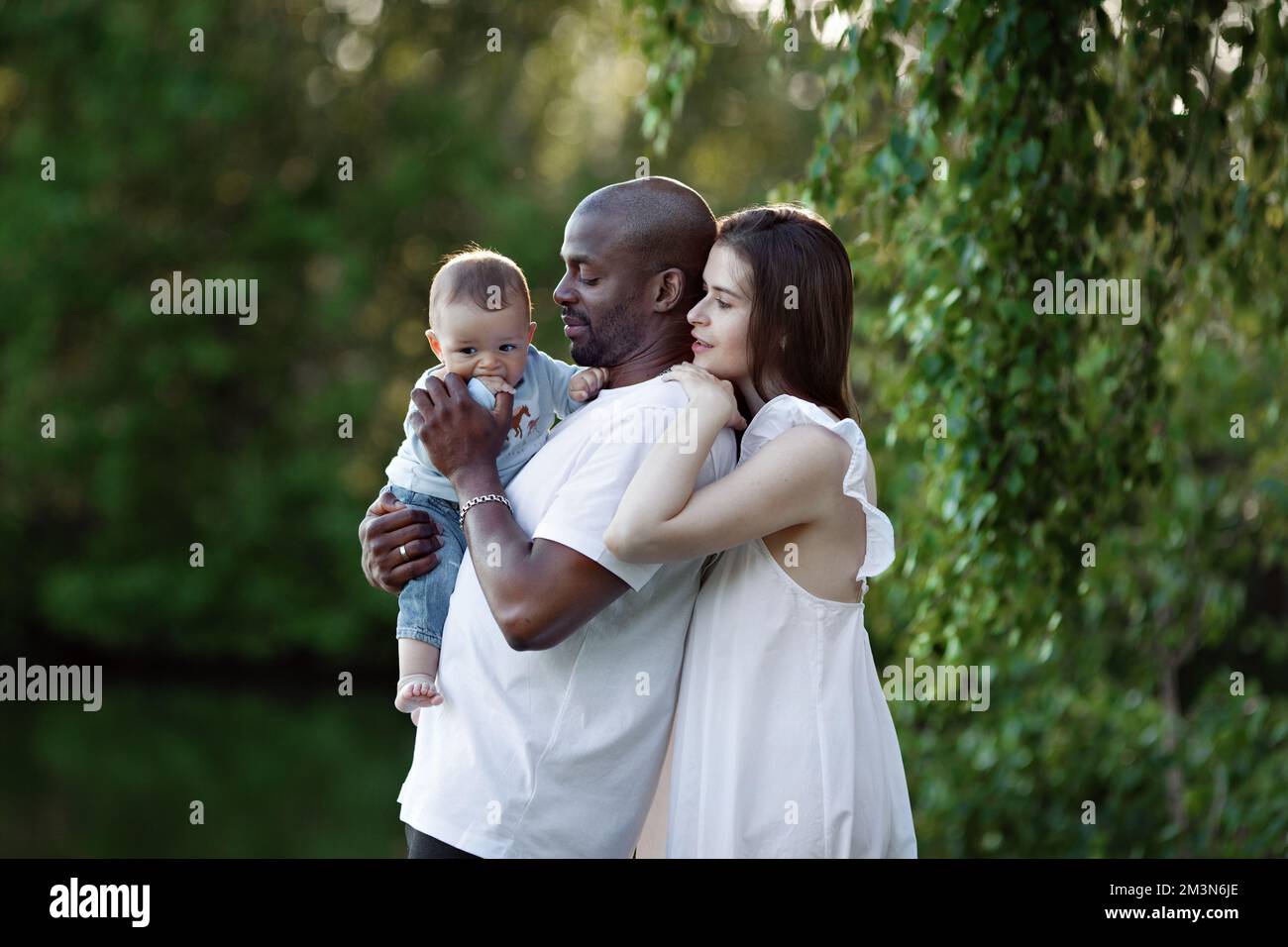 Happy young multiethnic family. Mother and father hugging baby. Parents, Portrait of Mom, dad and child on hands over green nature background Stock Photo