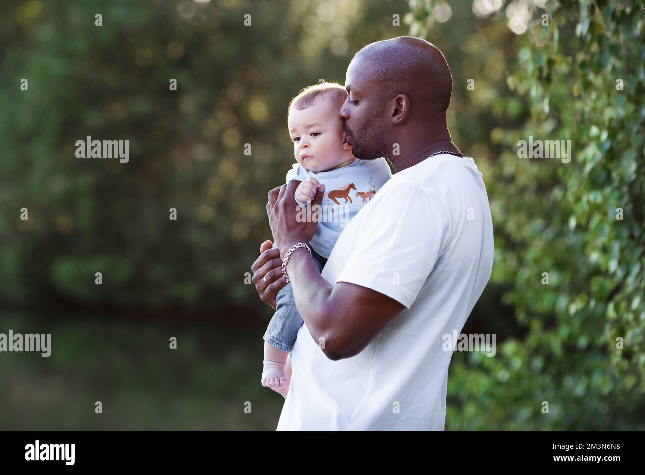 Father and son. African American dad kisses, holds on arms and loves baby multiethnic child against background of green nature Stock Photo