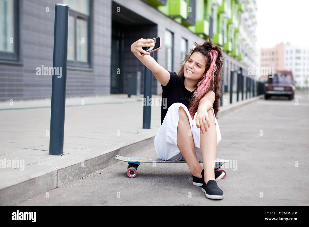 Teen girl sitting on road on skateboard, taking selfie on her phone and smiling. Lifestyle youth culture. Trendy teen with long pink colored hair Stock Photo