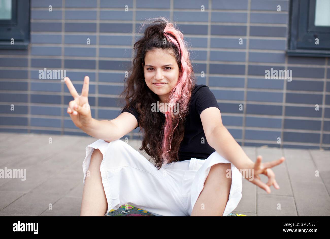 Portrait teen girl with long dark hair dyed pink, looking in camera and shows peace sign by hand on grey background Stock Photo