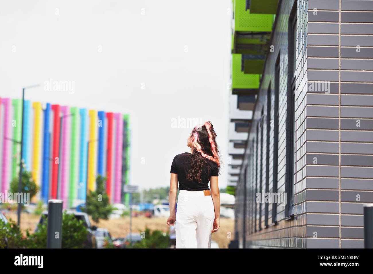 Back of unrecognizable girl with long hair dyed pink, walking along city street, against background of rainbow building Stock Photo