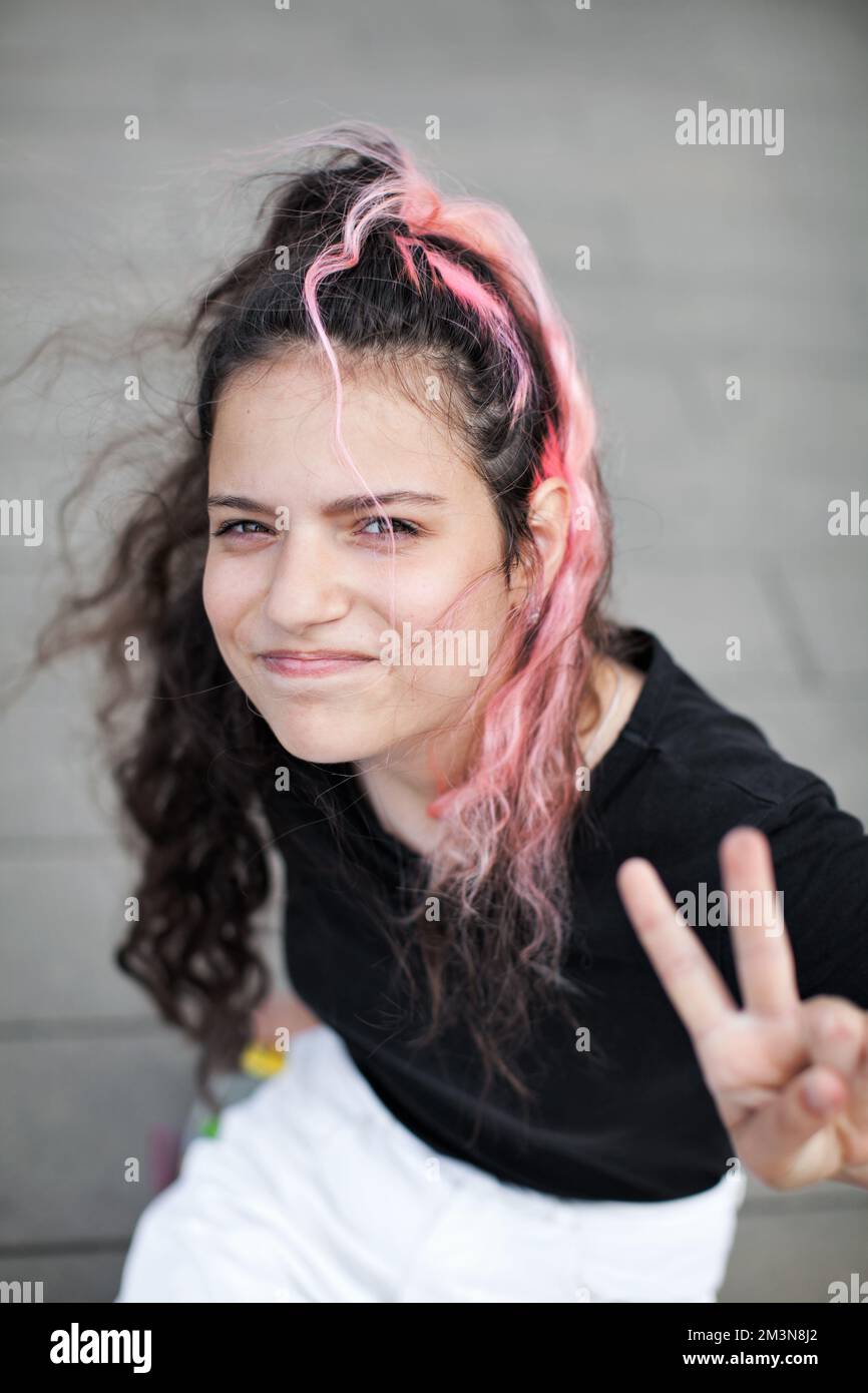 Portrait teen girl with long dark hair dyed pink, looking in camera and shows peace sign by hand on grey blurred background Stock Photo