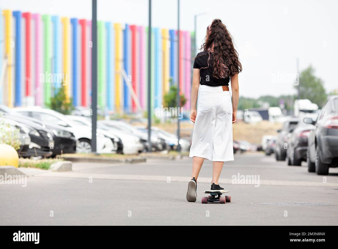 Back of unrecognizable girl, riding on skateboard along city street, against background of rainbow building Stock Photo
