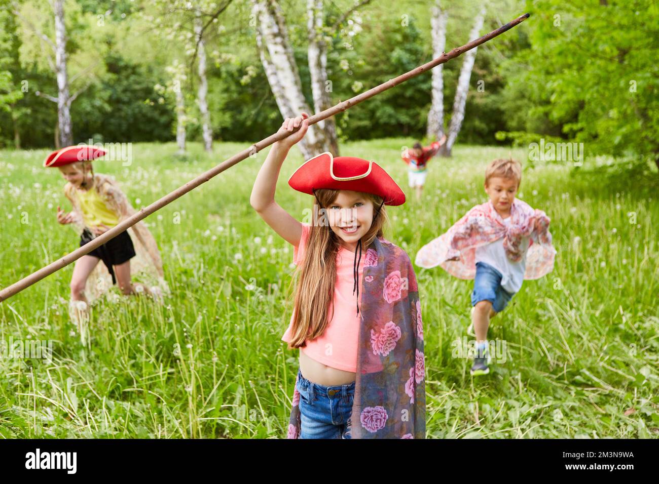 Portrait of smiling girl with tricorn hat and stick standing in grass by friends at park Stock Photo