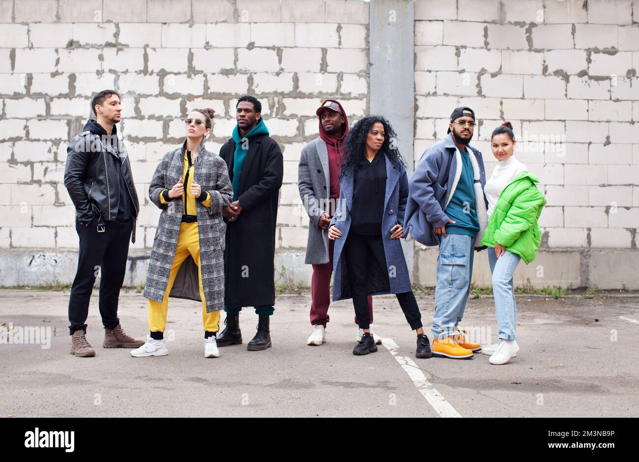 Multiethnic group of young hipsters stands together on street against gray brick wall. Diverse Stylish people in outerwear outdoor, youth culture Stock Photo