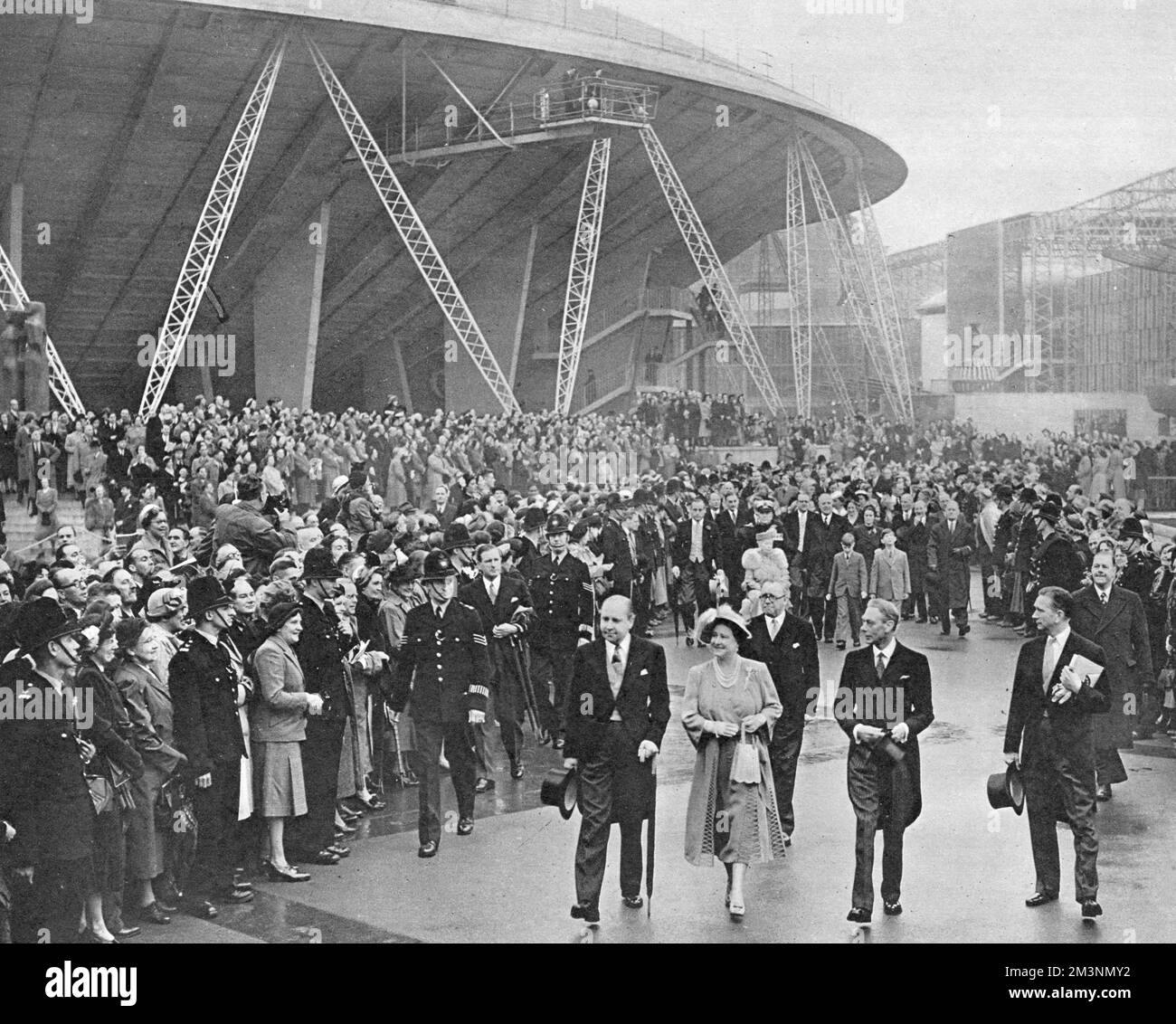 Scene at a royal visit to the Festival of Britain site on the South Bank, London.  Here King George VI and Queen Elizabeth are escorted by Gerald Barry, Director-General of the festival, followed by Herbert Morrison, Queen Mary (in a wheelchair), and other members of the royal family, watched by a large crowd of people.  The large building towering above them is the Dome of Discovery.        Date: 4 May 1951 Stock Photo