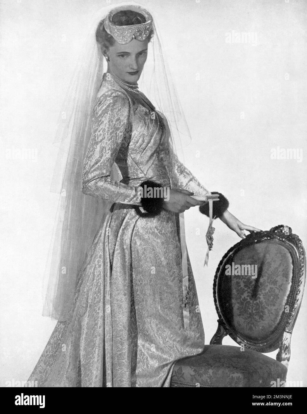 A wedding dress of pale gold and ivory patterned brocade with a prettily folded bodice and a train cut in one with the skirt.  The long, tight-fitting sleeves are cuffed with mink.     Date: 1953 Stock Photo