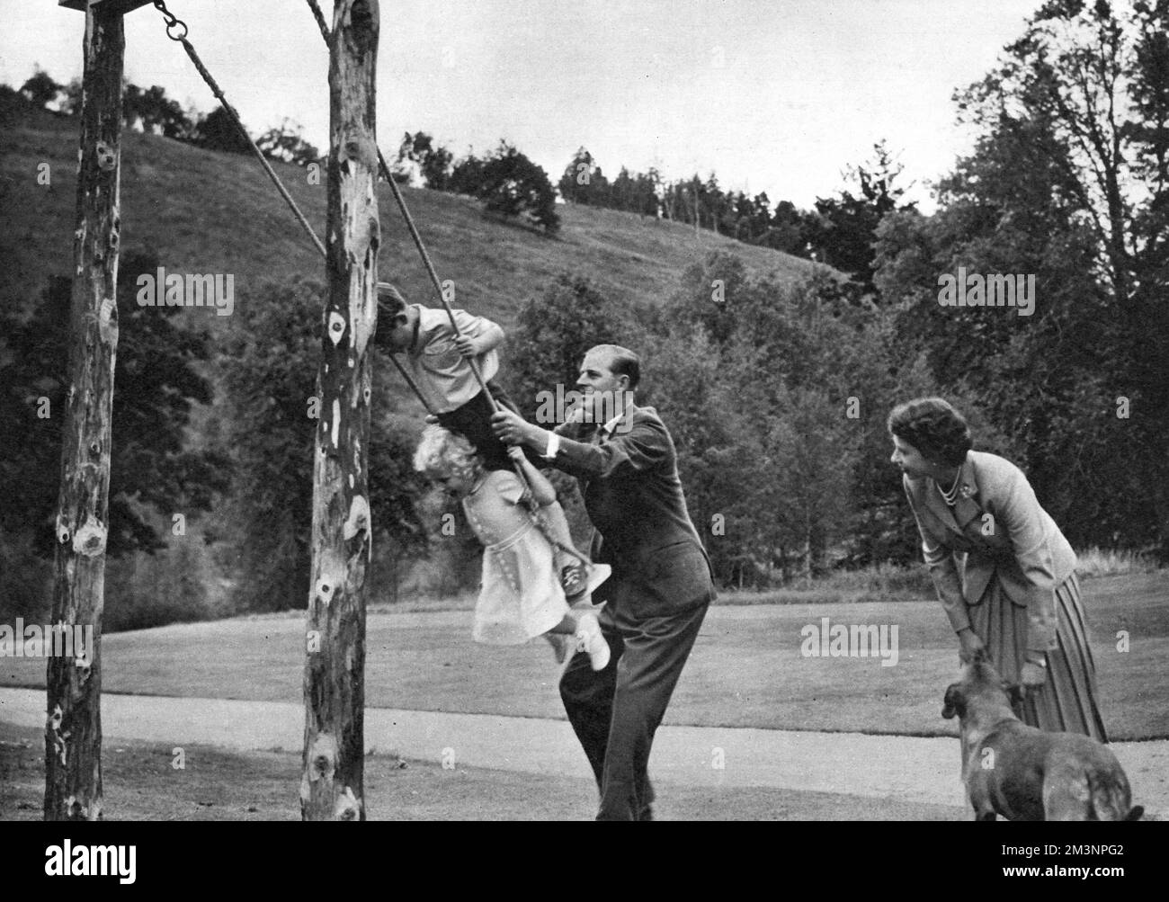 The Duke of Edinburgh pushes the young Duke of Cornwall and Princess Anne on a swing in the grounds of the royal residence at Balmoral. The Queen looks on with their dog, Candy.      Date: 1955 Stock Photo