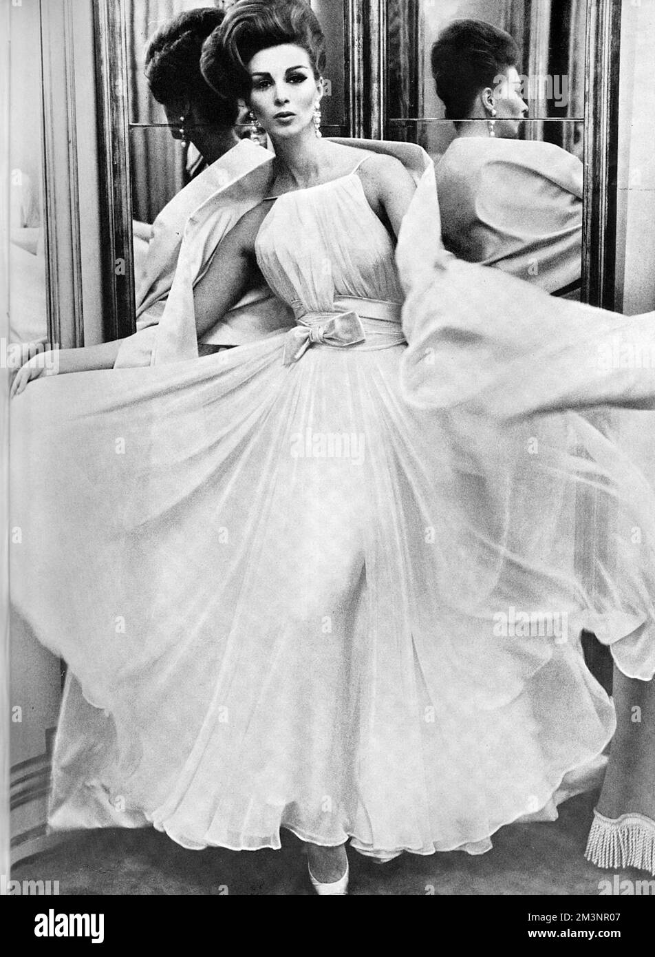 The return of the ballgown 'a la Grecque'; a pearly white chiffon dress with flounced skirt and shoestring strapped bodice swathed at the waist and accentuated by a bow of pink satin, worn with chandalier earrings in pearl and diamond.  By Balmain.     Date: 1962 Stock Photo