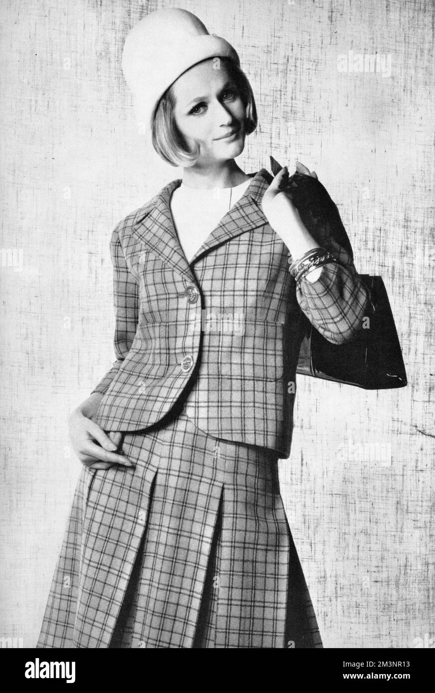 A box pleat skirt in blue plaid wool worn with a matching jacket and a white felt cloche hat and white sleeveless blouse, by Belinda Belville and worn by a model who looks remarkably like Glenn Close.  The bangles are by Christian Dior.     Date: 1962 Stock Photo