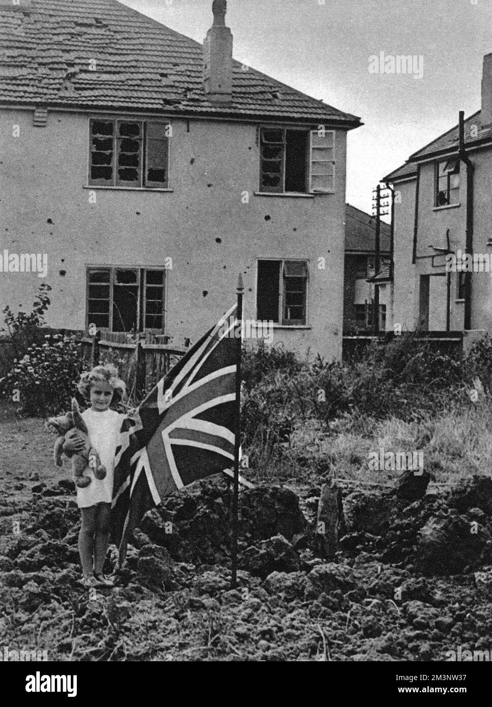 Defiant against German air raids, a little girl stands in her garden in Eastbourne, teddy bear in one hand, union jack in the other, posing by a bomb crater. Behind her is a house with smashed windows and missing slates: testimony to the violence of the explosion.     Date: 1940 Stock Photo