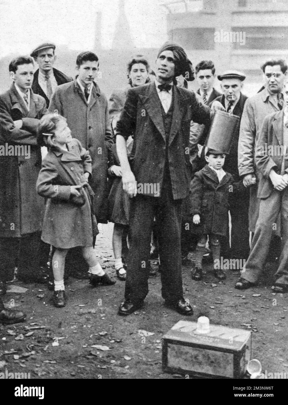 At Petticoat Lane, the London market, a young girl gazes up at a street performer wearing a turban who appears to be in the middle of a conjuring trick. The rest of the onlookers don't look quite so impressed.     Date: 1945 Stock Photo