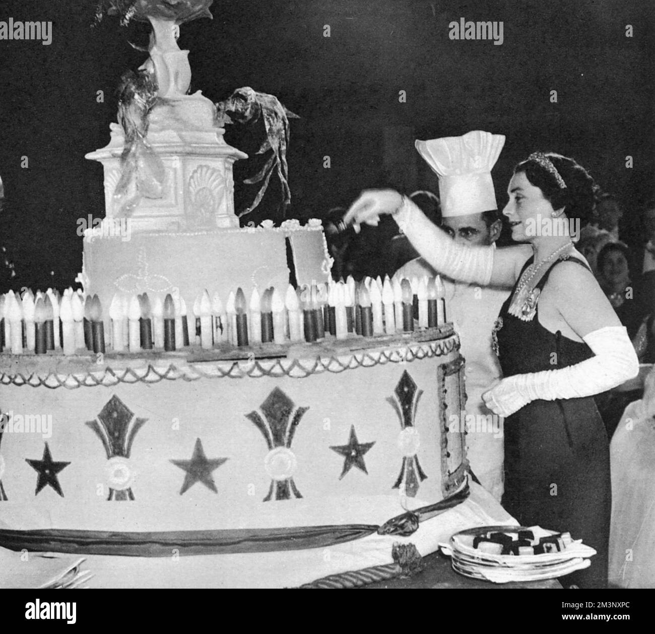 The Duchess of Gloucester (1901 -2004) cutting the giant cake holding 194 electric candles representing the number of years since the birth of Queen Charlotte at the ball named after her.  Queen Charlotte's Ball, held in honour of the wife of King George III and revived in 1925 to raise funds for the maternity hospital of the same name was one of the highlights of the London Season for a debutante.  All the girls would be dressed in white, and would dip into a carefully choreographed curtsy as the cake was cut.  There was apparently quite a scrum to get a piece of the cake afterwards!     Date Stock Photo