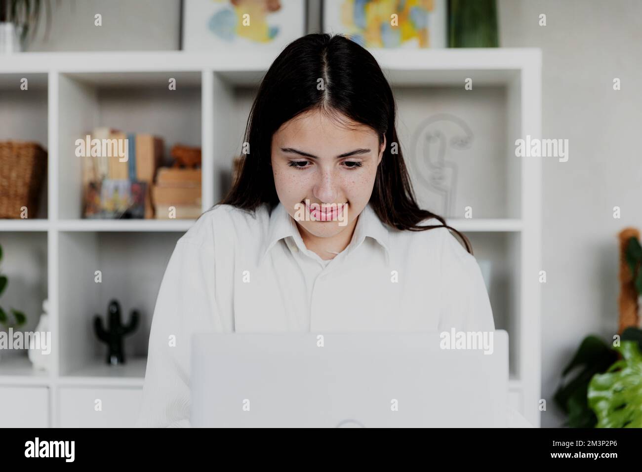 Brunette woman working with laptop at home or office. Girl is reading computer screen and smiling. Restrained joyful face. Blurred background with furniture. Person working from home concept Stock Photo