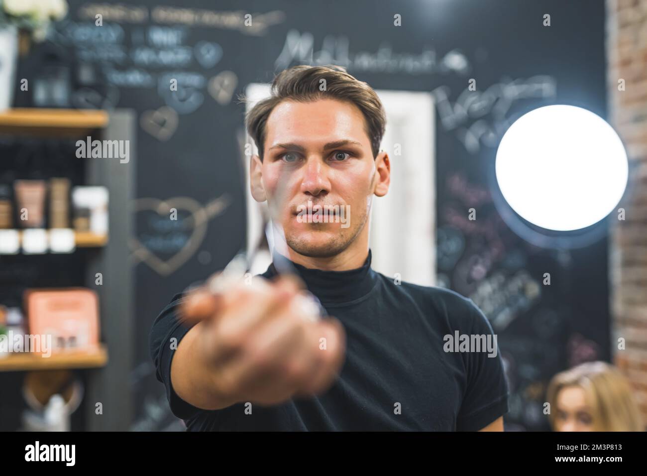 Professional focused male hairdresser in the black t-shirt showing blurred scissors to the camera in the professional hair salon. Blonde female client sitting in the background. High quality photo Stock Photo