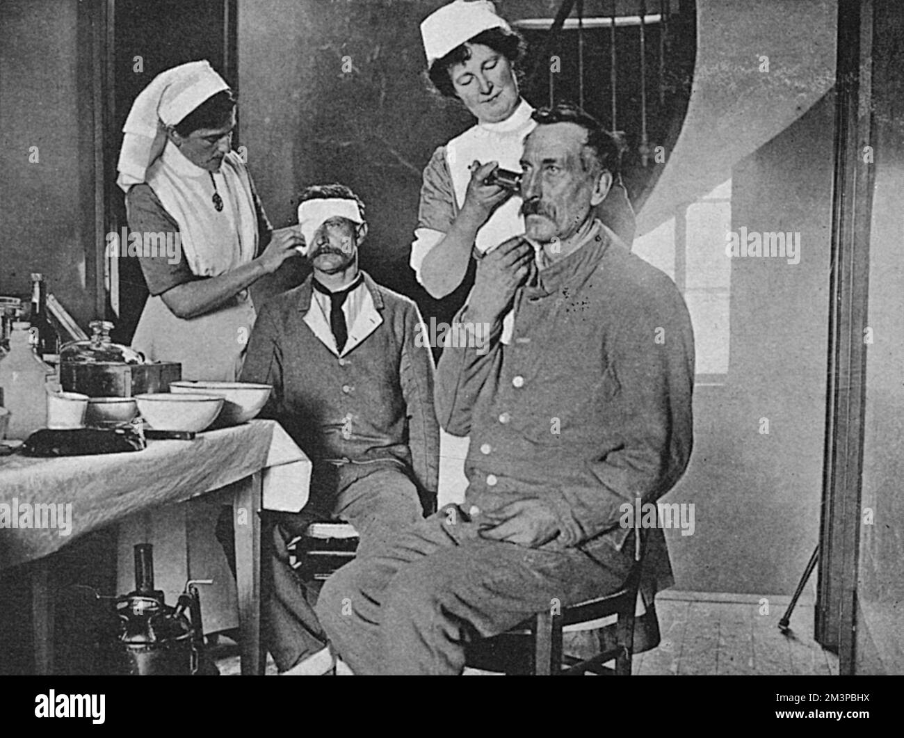 Two British nurses attached to the St. John's Voluntary Aid Detachment at attend to two French soldiers.  One is applying a dressing to the soldier's eye whilst the other is syringing antiseptic into the ear of a second soldier.     Date: 1916 Stock Photo