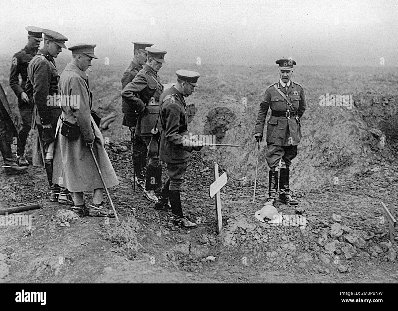 King George V beside the grave of a British soldier, marked with a cross and a steel helmet, on the battlefield during a visit to the Front in 1916. Among the others accompanying the King is Edward, Prince of Wales (later King Edward VIII, Duke of Windsor), who is seen in a great coat smoking a cigarette.     Date: 1916 Stock Photo