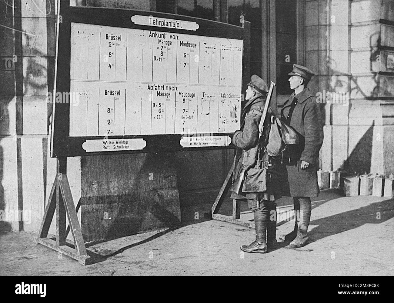 Two British soldiers stand looking at the railway timetable -written in German -  at Mons station at the end of the First World War.  Places such as Tournai and Valenciennes, which had been captured by the Allies had been removed from the board.     Date: 1918 Stock Photo