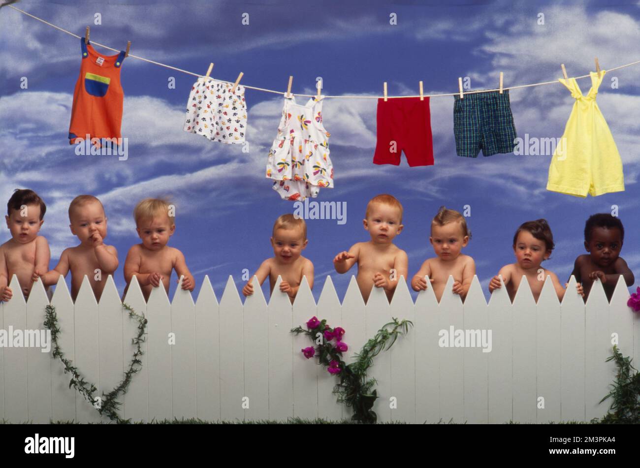 Row of naked babies standing behind a white picket fence with laundry