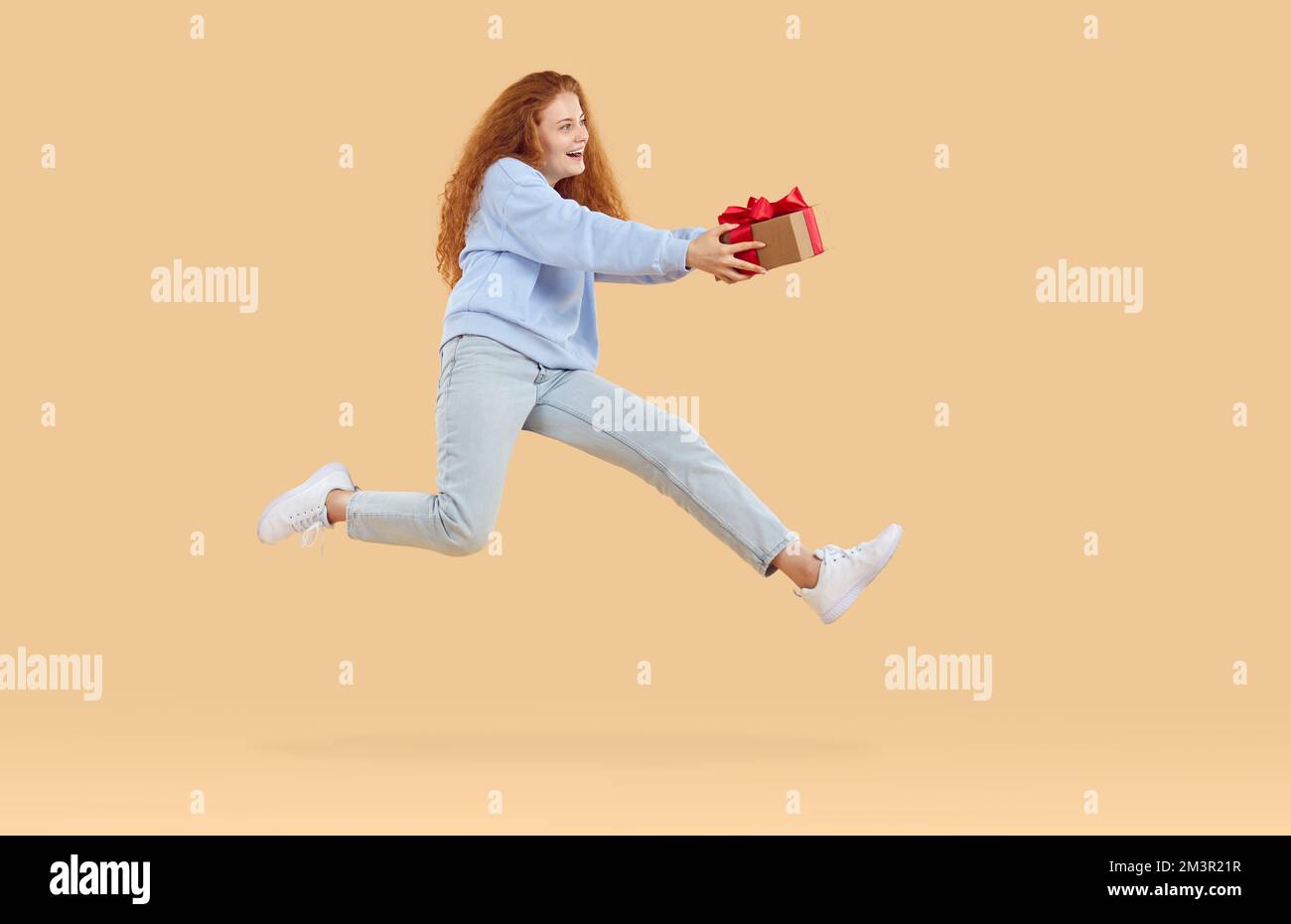 Happy curly redhead girl jumping in air holding Xmas gift present in hands on beige background. Stock Photo