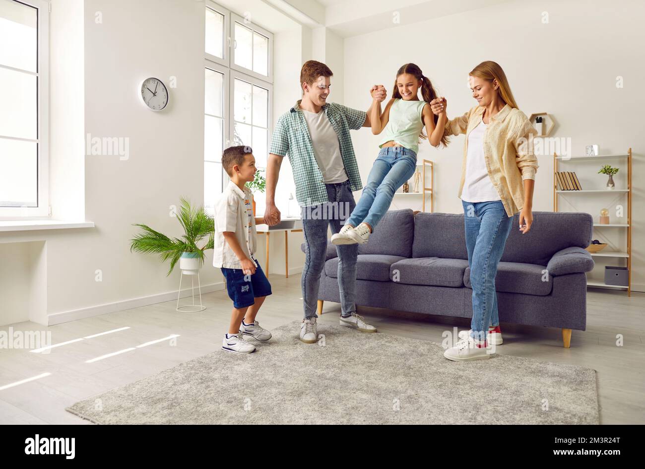 Happy family are playing together, girl is jumping from sofa holding hands parents in living room. Stock Photo