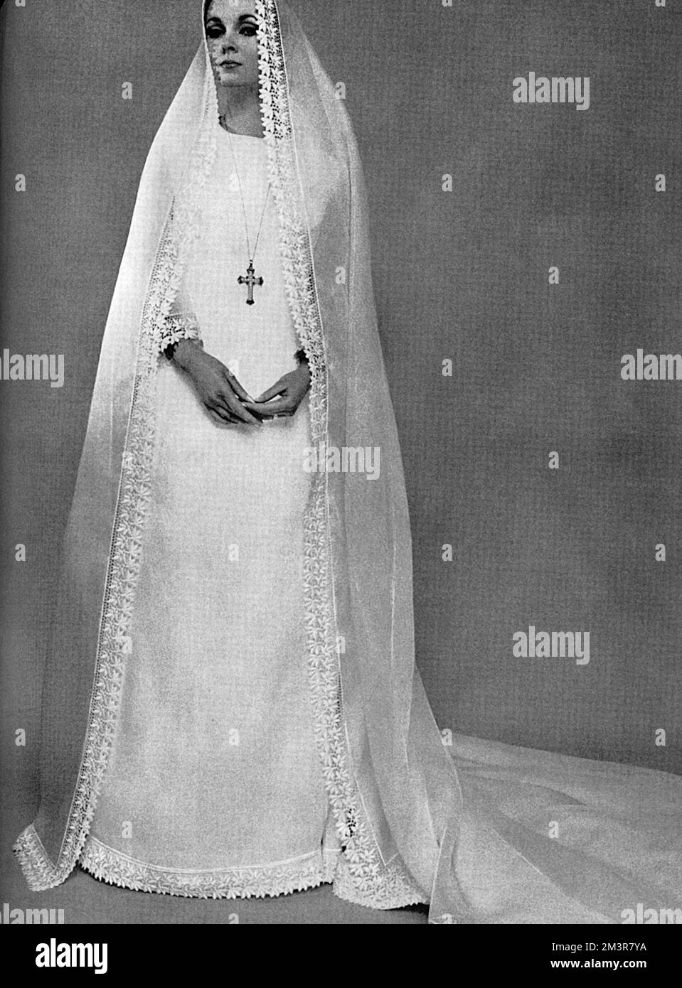 A Camelot-style wedding dress and veil of nun-like simplicity in translucent white organdie with a wide border of guipure lace and the cuffs and hem, also trimming the veil and train.  By Belinda Belville.     Date: 1965 Stock Photo