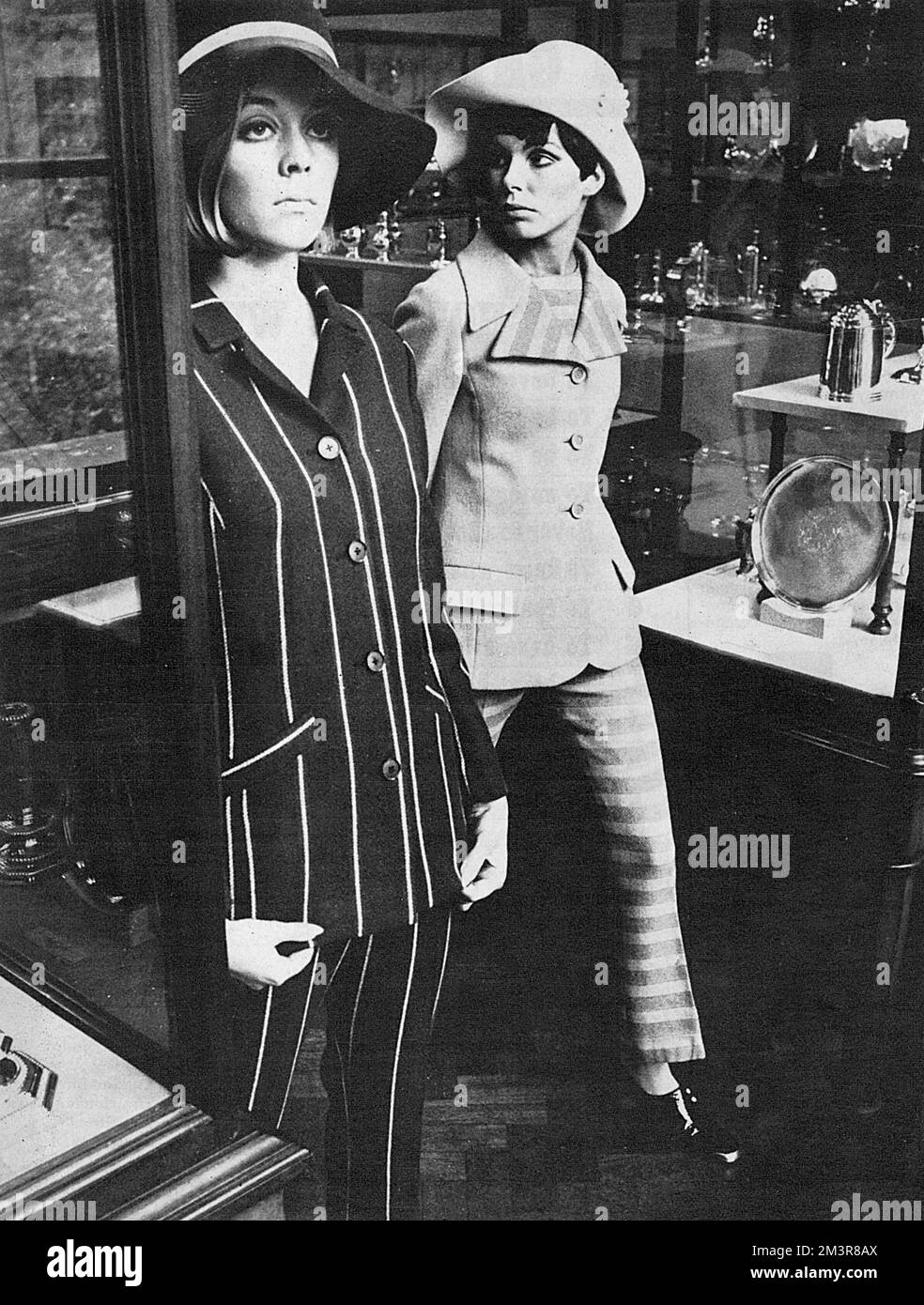 Model on the left wears a navy with red and white striped Mono trouser suit, with Edward Mann 'spy girl hat' from Bourne and Hollingsworth. Model on the right wears a shocking-pink trouser suit with striped trousers and sleeveless top by Carol Freedman and pink hat with white daisy from Herbert Johnson.     Date: 1966 Stock Photo