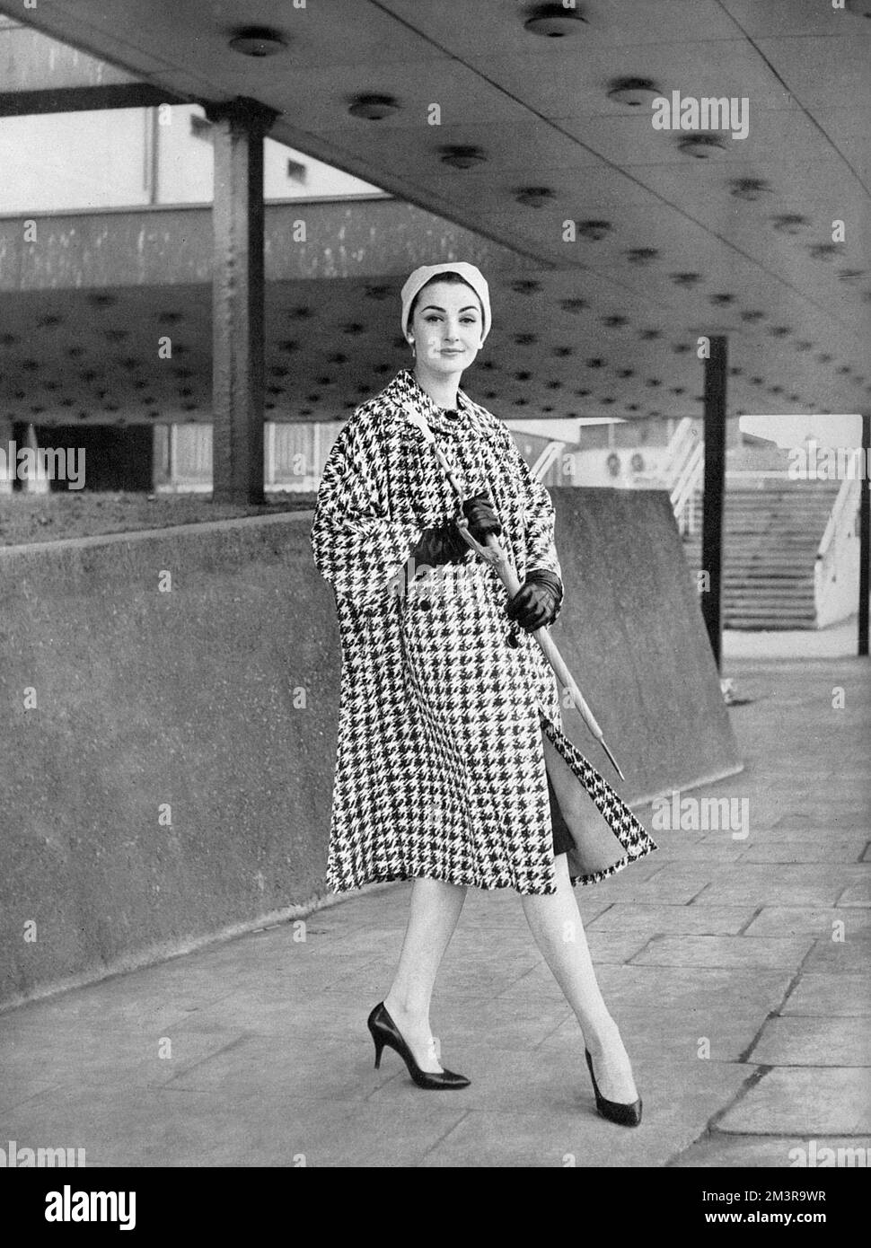 A Burberry in French cotton printed in large black and white houndstooth design, fully shower proof and lined with cherry taffeta.  The collarless neckline and three quarter length sleeves make it a good choice for town.  Worn with a white-proofed turban from Simone Mirman and an umbrella from James Smith and Son.       Date: 1959 Stock Photo