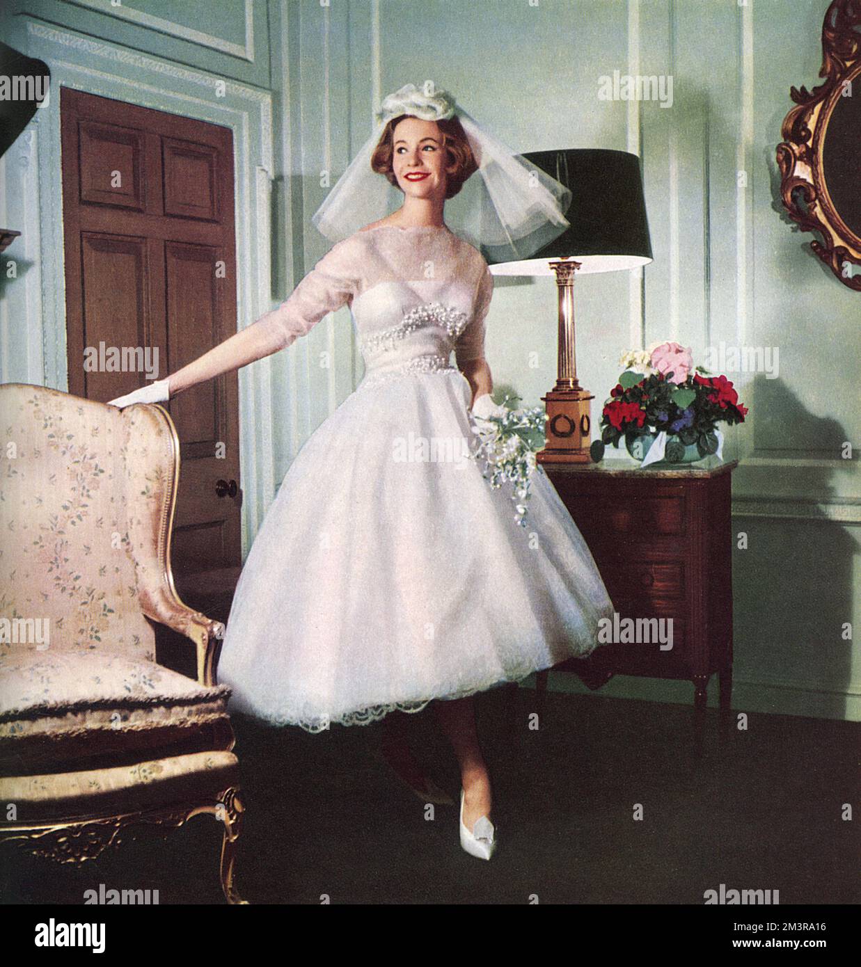 A high fashion wedding dress in lace tinted with the merest pink, short enough in front to show a stocking and slippered foot.  By Carina Martin from Harrods Trousseau room worn with an organza rose headdress and satin Dior shoes.       Date: 1958 Stock Photo