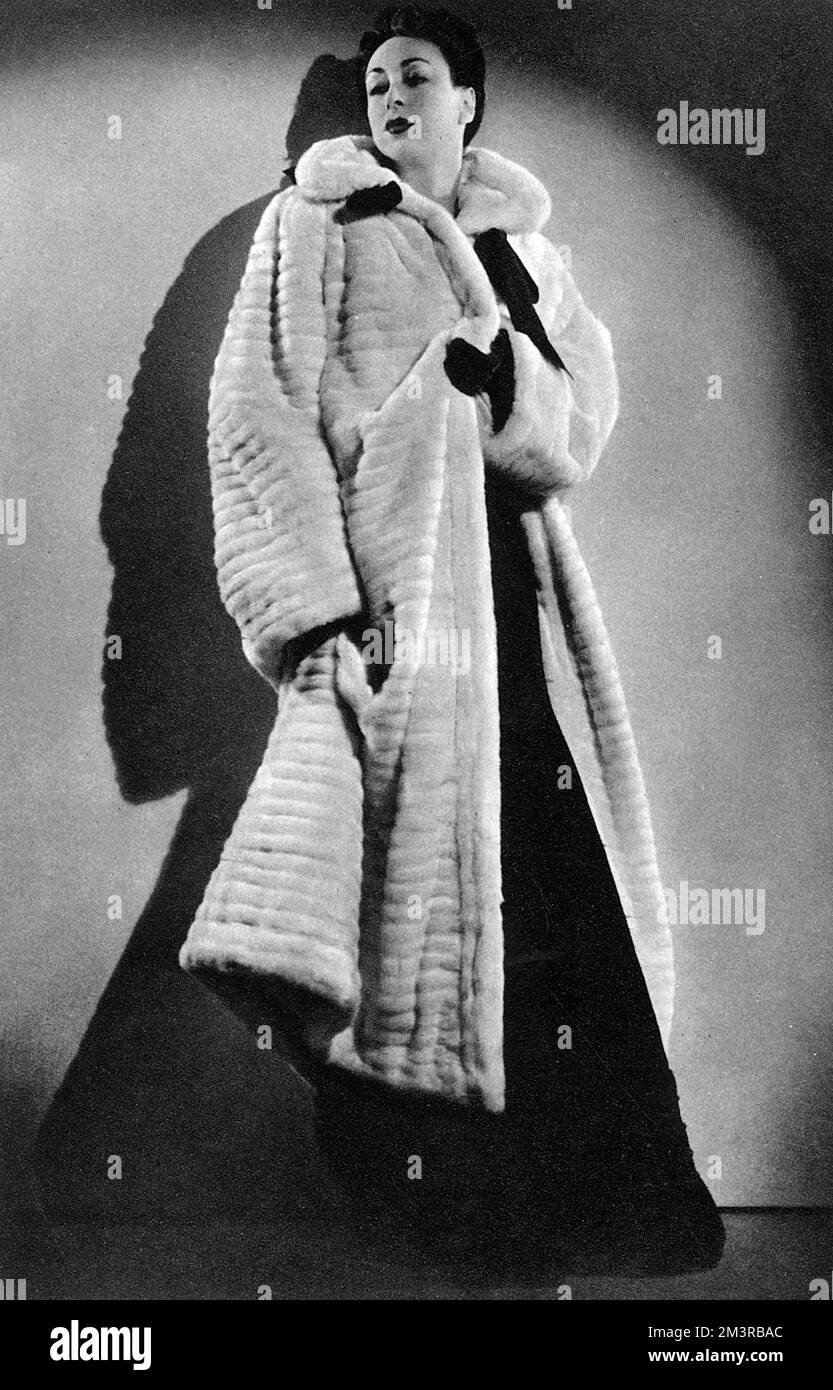 Photograph of a model wearing a white ermine coat designed by Elsa Schiaparelli. The fur is worked horizontally, the coat falls in loose folds from gathered shoulders and is worn with violet gloves over a violet velvet dress.  1937 Stock Photo