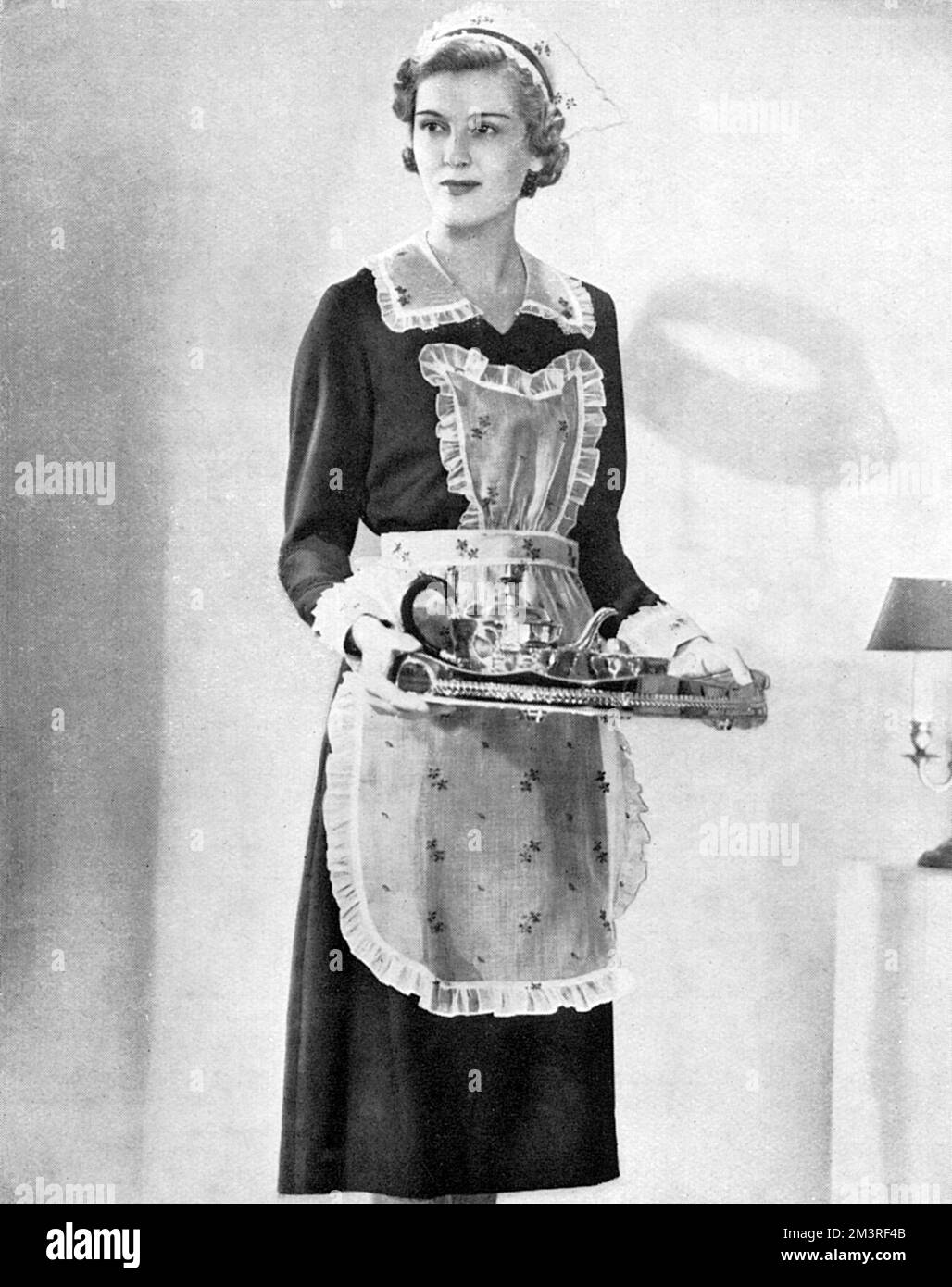 Advertisement for Garrould's of Edgware Road, London, highlighting their uniforms for staff.  'Distinguished hostesses who take a personal interest in their entertaining to express their artistry and good taste, select uniforms for their staff at Garrould's'.  The uniform shown comprises a white organdie apron with a wine blue or green embroidered sprig, cap to match with two goffered frills at top and bottom, velvet ribbon to match and collar and cuffs to match.       Date: 1938 Stock Photo