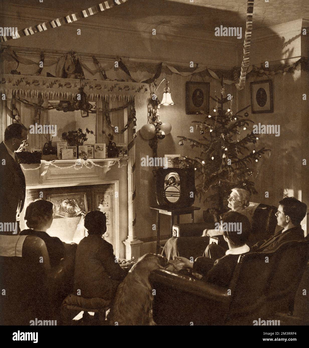 A 1950s family sit around the fire at Christmas time listening to a wireless broadcast  Streamers and paper chains hang from the ceiling and a rather mean looking Christmas tree stands in the corner.        Date: 1952 Stock Photo