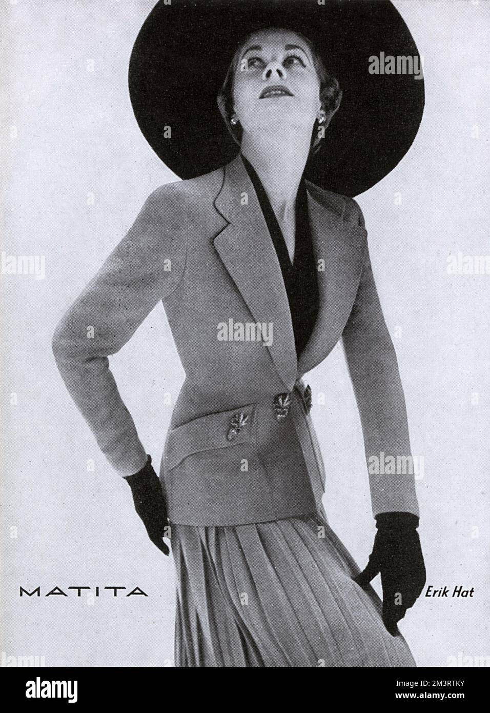 Advertisement for clothing brand, Matita featuring a model wearing a tailored, fitted jacket with a matching pleated skirt and a hat by Erik.    1950 Stock Photo